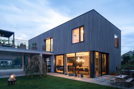 Modernes Holzhaus mit großen Fenstern und Gartenbeleuchtung bei Dämmerung.