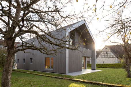 Modernes graues Haus mit schrägem Dach, umgeben von Bäumen und einem Garten.